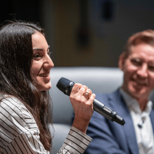 Gabriela Hersham, Regent's alumna and founder of Huckletree, speaking using a microphone at an Alumni Association event. Geoff Smith, CEO and Vice-Chancellor of Regent's University London, is in the background.